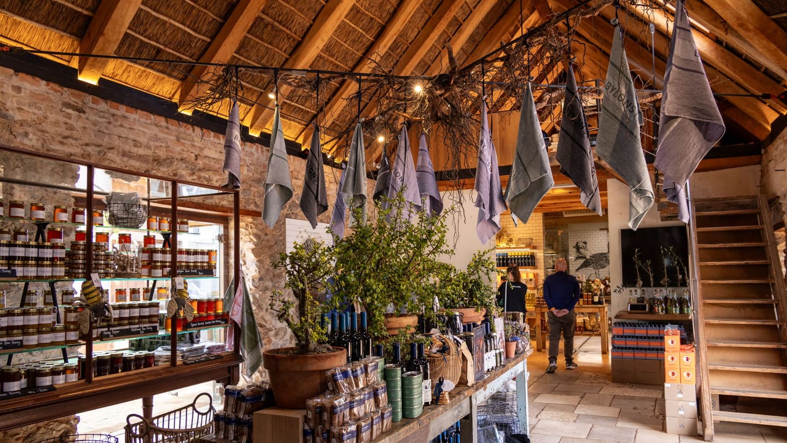 Inside a rustic farm shop with stone walls and timber beams, shelves of jars, wine and local produce in warm light